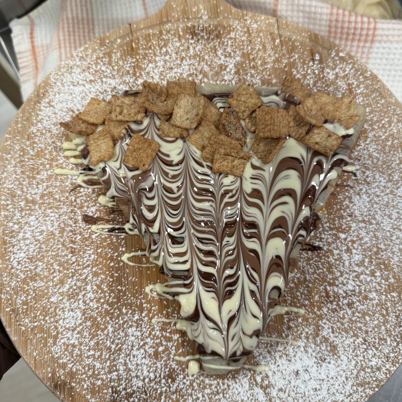 Cinnamon Toast Crunch Crêpe with a chocolate and white chocolate swirl, topped with Cinnamon Toast Crunch cereal on a board dusted with powdered sugar