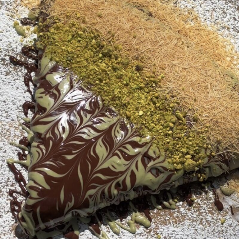 Dubai Crêpe with a chocolate and pistachio swirl, crushed pistachios, and toasted kataifi on a board dusted with powdered sugar